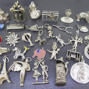 May include: A collection of silver charms in various shapes, including a theater mask, Eiffel Tower, and a beer stein. Other charms depict animals, figures, and a map of Belgium. The charms are displayed on a dark surface.