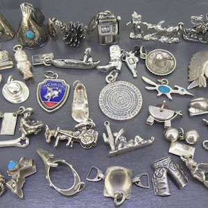 May include: A collection of silver charms and pendants, including a slot machine, cowboy boots, a sombrero, and a Native American headdress. Other charms include a shoe, a Nevada shield, and a rope. The charms are displayed on a dark grey surface.