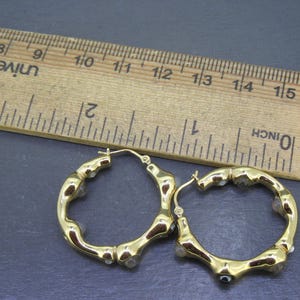 May include: A pair of gold-colored hoop earrings with a bamboo-like design, accented with small, clear gemstones. A wooden ruler with inch measurements is in the background.