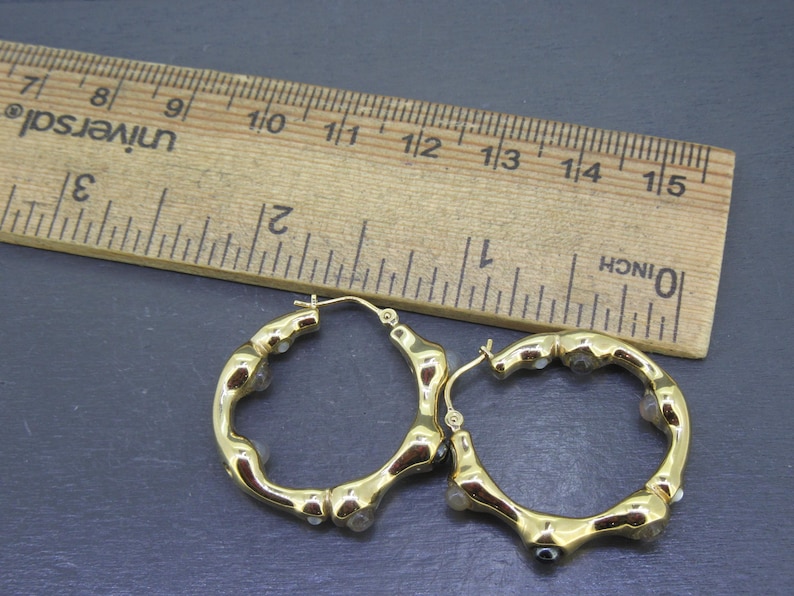 May include: A pair of gold-colored hoop earrings with a bamboo-like design, accented with small, clear gemstones. A wooden ruler with inch measurements is in the background.