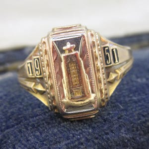 May include: A gold-toned class ring with the year 1951 displayed on the sides. The ring features a central rectangular design with an embossed building and a star-like emblem. The ring is set against a blue fabric background.