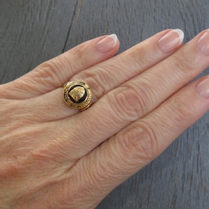 May include: A gold ring featuring a circular black inlay with a central gold emblem. The ring band has a textured, decorative border surrounding the black center piece, creating a distinct contrast between the gold metal and the dark inlay.