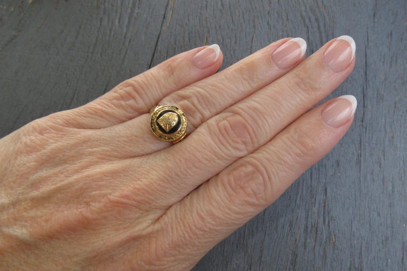 May include: A gold ring featuring a circular black inlay with a central gold emblem. The ring band has a textured, decorative border surrounding the black center piece, creating a distinct contrast between the gold metal and the dark inlay.