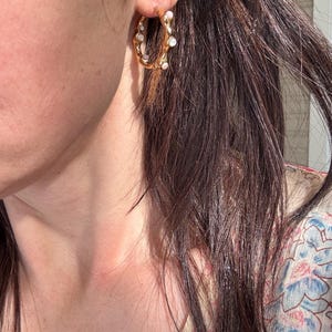 May include: Gold-tone hoop earrings with irregular, textured design. The hoops are adorned with small, white, pearl-like embellishments. The earrings are shown on a person's ear, with dark hair in the background.