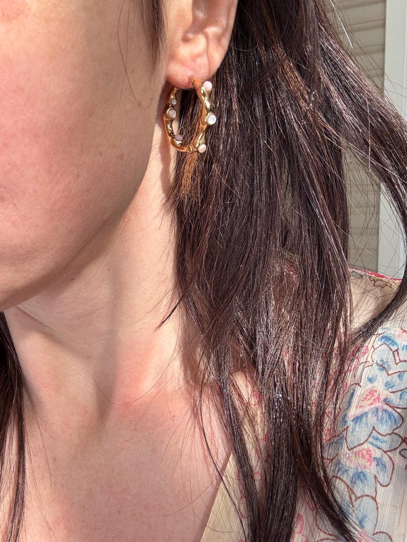 May include: Gold-tone hoop earrings with irregular, textured design. The hoops are adorned with small, white, pearl-like embellishments. The earrings are shown on a person's ear, with dark hair in the background.