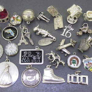 Può includere: Una collezione di ciondoli vintage in argento, tra cui un ditale, un pallone da basket, la Torre Eiffel, un aereo e un ciondolo "Sweet 16". Altri ciondoli raffigurano un orologio, una lavagna scolastica e uno stivale. I ciondoli sono su una superficie grigio scuro.