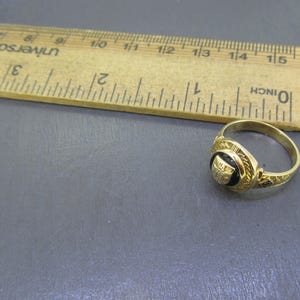May include: A gold ring with a circular black inlay featuring a small gold emblem, positioned next to a wooden ruler that shows the ring has a diameter of approximately 0.75 inches. The ring band has a textured, patterned design.