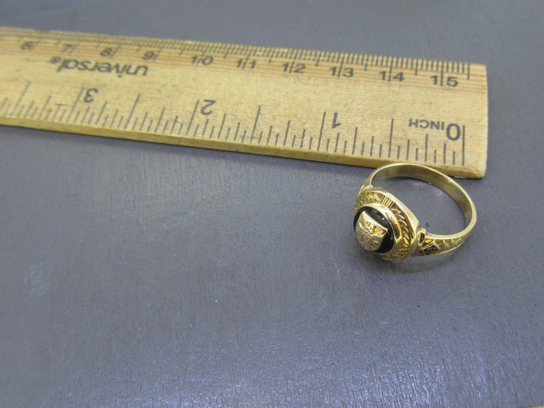 May include: A gold ring with a circular black inlay featuring a small gold emblem, positioned next to a wooden ruler that shows the ring has a diameter of approximately 0.75 inches. The ring band has a textured, patterned design.