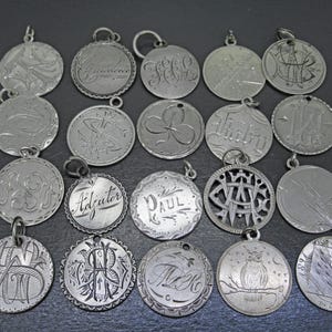 May include: A collection of twenty circular silver-toned metal charms arranged in four rows. Each charm features unique engraved designs, including monograms, script lettering such as 'Adjutor' and 'Paul', a clover, an owl, and geometric patterns.