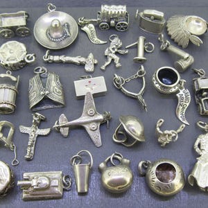 May include: A collection of silver-toned charms, including a sombrero, airplane, well, and a Native American headdress. Other charms include a tank, a bucket, and a miniature wagon. The charms are displayed on a dark gray surface.