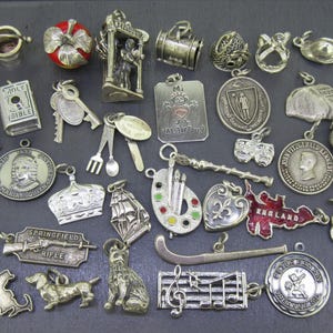 Pode incluir: Uma coleção de amuletos e pingentes prateados, incluindo um livro em miniatura, chaves, uma coroa, um navio e um dachshund. Alguns amuletos apresentam texto, como "SPRINGFIELD RIFLE" e "MASS."