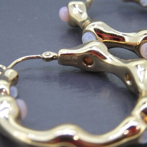 May include: Close-up of gold-toned hoop earrings with a unique, textured design. Each earring features small, light pink gemstones. The earrings are marked "14K FP China".