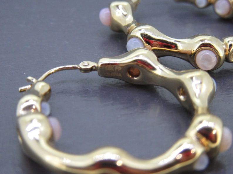 May include: Close-up of gold-toned hoop earrings with a unique, textured design. Each earring features small, light pink gemstones. The earrings are marked "14K FP China".