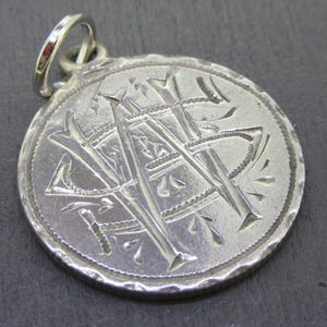May include: A silver pendant with a circular design. The pendant features an engraved monogram of two intertwined letters, possibly initials, within a decorative border. A small loop is attached at the top for a chain.