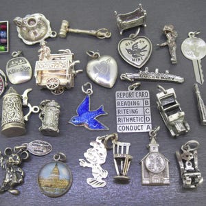 May include: A collection of silver charms, including a miniature church, a report card, a blue bird, and a miniature Hoover Dam sign. Other charms include a key, ballet slippers, and a heart-shaped charm with a bird design.