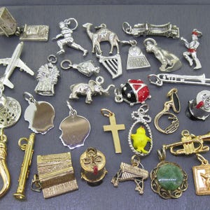 Könnte beinhalten: Eine Sammlung von silber- und goldfarbenen Charms in verschiedenen Formen, darunter ein Flugzeug, ein Kamel, ein Kreuz und ein Marienkäfer. Weitere Charms sind Musikinstrumente, eine Glocke und eine Profilsilhouette. Die Charms sind wahrscheinlich für ein Armband oder eine Halskette.