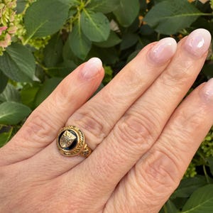 May include: A gold ring featuring a circular black onyx inlay centered with a gold crest emblem. The band has a textured, decorative border surrounding the central stone, showcasing intricate metalwork details on the shank.