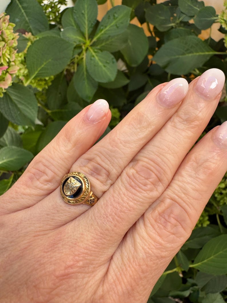 May include: A gold ring featuring a circular black onyx inlay centered with a gold crest emblem. The band has a textured, decorative border surrounding the central stone, showcasing intricate metalwork details on the shank.