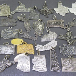 May include: A collection of vintage silver-toned state charms, each shaped like a state outline. Some charms have engraved details and text, including "KANSAS", "MASS", "CALIFORNIA", and "COLORADO". The charms are on a dark gray surface.