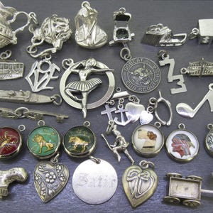Puede incluir: Una colección de dijes y colgantes de plata, que incluyen una bailarina, un tren, una llave, una cruz y un emblema de los Boy Scouts. Algunos dijes tienen detalles de esmalte de colores. Los dijes se muestran sobre una superficie oscura.