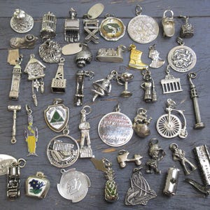 Op de afbeelding: Een verzameling vintage zilveren bedels in diverse vormen, waaronder een bel, een vogelkooi, een stoel, een laars en een fiets. Sommige bedels hebben emaille details. De bedels worden getoond op een donkergrijze houten ondergrond.