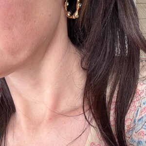 May include: Gold-toned hoop earrings with an organic, textured design. The earrings are adorned with small, clear and dark-colored stones. The jewelry is a stylish accessory, suitable for various occasions.