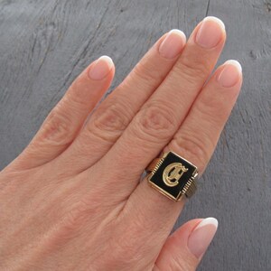 May include: A gold-toned ring with a black square face featuring a gold letter "C". The ring has a ribbed band and a rectangular black center. The ring is on a person's finger.