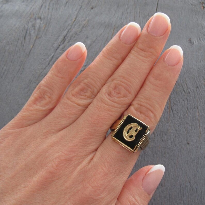 May include: A gold-toned ring with a black square face featuring a gold letter "C". The ring has a ribbed band and a rectangular black center. The ring is on a person's finger.