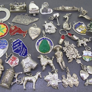 May include: A collection of vintage silver charms, including a sewing machine, piano, and state-shaped pendants. Other charms include a shoe, a cuckoo clock, and a heart-shaped charm with "Happy Birthday" text. The charms are on a dark gray surface.