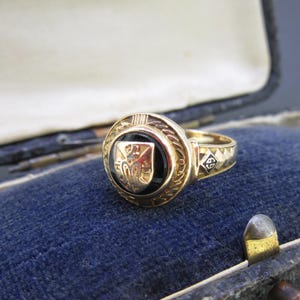 May include: A gold ring featuring a circular black onyx inlay centered with a gold shield emblem. The band displays a textured pattern and a small square detail with the number 69 engraved in black. The ring has a polished metallic finish.