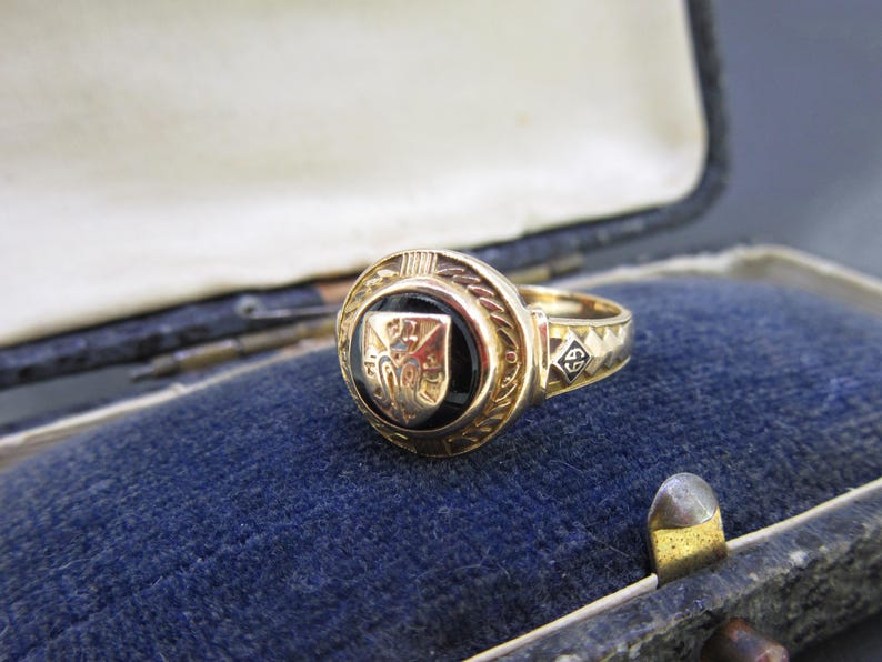 May include: A gold ring featuring a circular black onyx inlay centered with a gold shield emblem. The band displays a textured pattern and a small square detail with the number 69 engraved in black. The ring has a polished metallic finish.