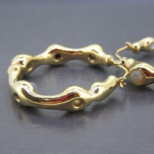May include: Close-up of two gold-colored hoop earrings. The earrings have an organic, textured design with a series of rounded shapes and small holes. The earrings are on a gray surface. The earrings are a jewelry item.