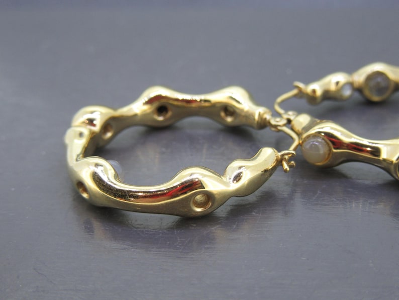 May include: Close-up of two gold-colored hoop earrings. The earrings have an organic, textured design with a series of rounded shapes and small holes. The earrings are on a gray surface. The earrings are a jewelry item.