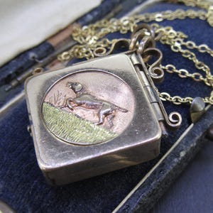 May include: A gold-toned square pendant necklace with a detailed dog illustration on a circular face. The dog is depicted in a field of green grass. The pendant is on a gold chain and rests on a blue velvet surface.