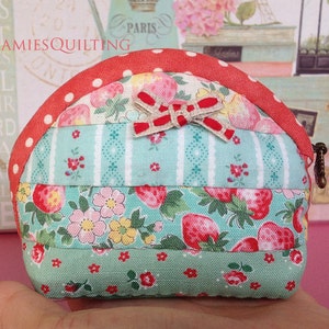May include: A small, quilted coin purse with a red, white, and green patchwork design. The purse has a red and white polka dot trim and a bow made of red and white ribbon. The text "JAMIESQUILTING" is visible on the purse.
