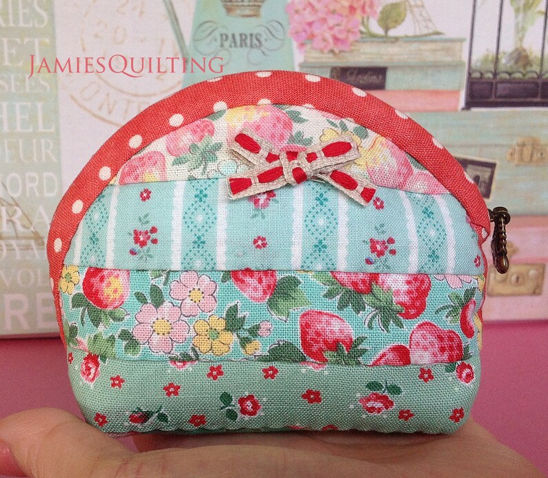 May include: A small, quilted coin purse with a red, white, and green patchwork design. The purse has a red and white polka dot trim and a bow made of red and white ribbon. The text "JAMIESQUILTING" is visible on the purse.