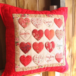 Op de afbeelding: Rood gestikt kussen met een crèmekleurig paneel met een patchwork van harten met geborduurde namen, waaronder "Sarah", "Isabella", "Joshua" en "New Baby of 2014".