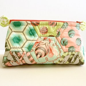 Peut inclure: Une pochette zippée en patchwork avec un motif hexagonal. Le tissu est un mélange de vert, rose et gris avec des motifs géométriques et floraux. La fermeture éclair est de couleur orange vif.