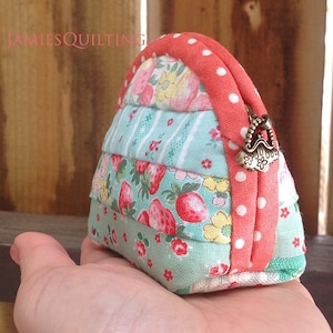 May include: A small, handmade coin purse with a patchwork design featuring floral and strawberry prints. The purse is made of a variety of fabrics in shades of blue, green, pink, and white. The purse has a coral-colored zipper with white polka dots and a brass clasp.