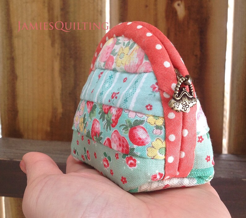 May include: A small, handmade coin purse with a patchwork design featuring floral and strawberry prints. The purse is made of a variety of fabrics in shades of blue, green, pink, and white. The purse has a coral-colored zipper with white polka dots and a brass clasp.