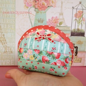 Puede incluir: Una pequeña cartera de monedas de tela de patchwork rojo, blanco y verde con un lazo y un cierre de cremallera.