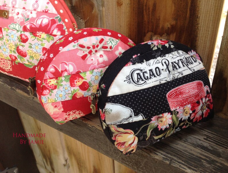 Może przedstawiać: Trzy ręcznie robione portmonetki z tkaniny w r&oacute;żnych wzorach. Pierwsza portmonetka jest czerwona i r&oacute;żowa z truskawkami i kwiatami. Druga portmonetka jest czerwona i biała z truskawkami i kwiatami. Trzecia portmonetka jest czarna w białe kropki i z kwiatowym wzorem. Portmonetka ma napis "Cacao-Payraud".