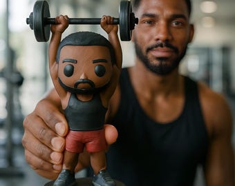 Puede incluir: Una figura de un levantador de pesas con una camiseta sin mangas negra y pantalones cortos rojos, levantando una barra. La figura tiene una tez oscura, barba y está sobre una base negra. La barra tiene pesas negras en cada extremo. El fondo está borroso.