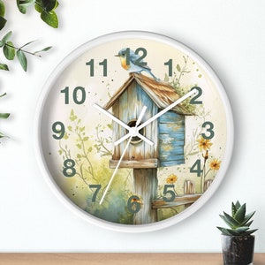 May include: White round clock with a bluebird perched on a birdhouse. The birdhouse is painted blue and white with a brown roof. The clock face has green numbers and a white background. The clock is surrounded by a white frame.