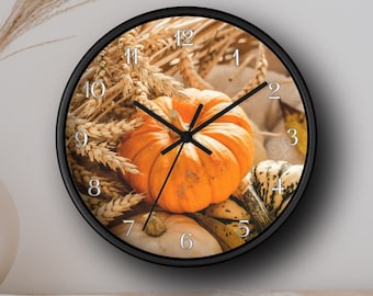 May include: A round wall clock with a black frame. The clock face features a still life of pumpkins and wheat stalks in shades of orange, yellow, and brown. Silver numerals mark the hours. The clock hands are black.
