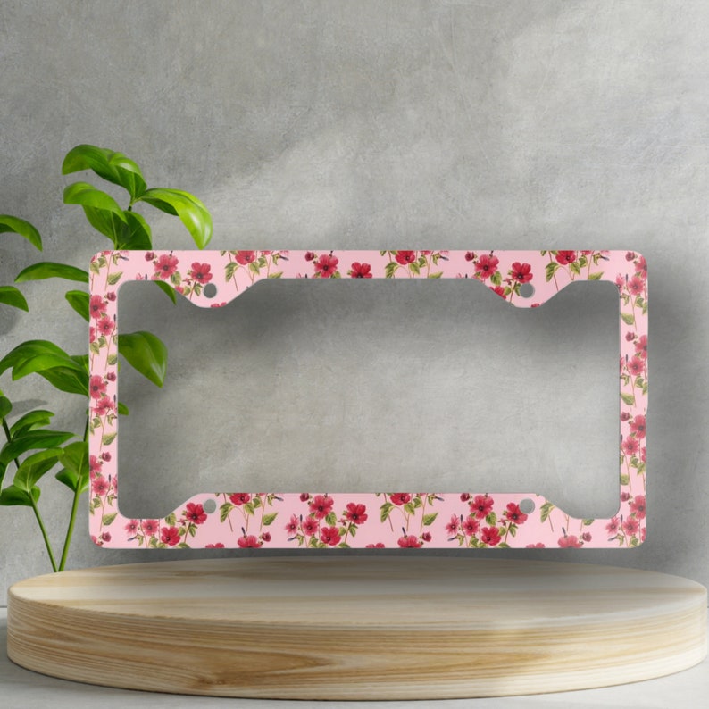 Op de afbeelding: Een roze kentekenplaathouder met een bloemenpatroon met rode bloemen en groene bladeren.