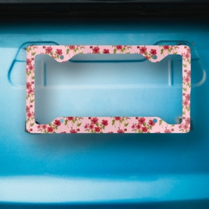 Op de afbeelding: Roze kentekenplaathouder met een bloemenpatroon met rode bloemen en groene bladeren.