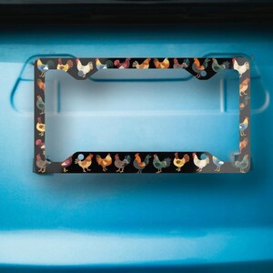 May include: Black license plate frame featuring a repeating pattern of colorful rooster illustrations. The roosters are depicted in various poses and colors, set against a black background. The frame is designed to fit a standard U.S. license plate.