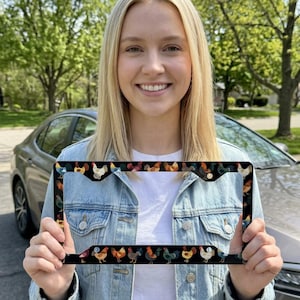 May include: Black license plate frame featuring a repeating pattern of colorful rooster illustrations. The frame is held up in front of a car and a person. The roosters are in various colors, including red, yellow, and blue.