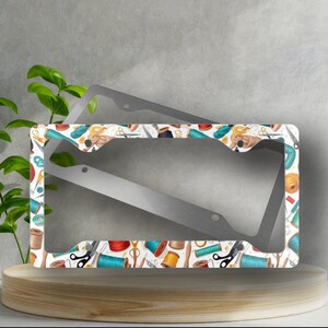 May include: A white metal license plate frame with a colorful pattern of sewing supplies, including spools of thread, scissors, and needles.